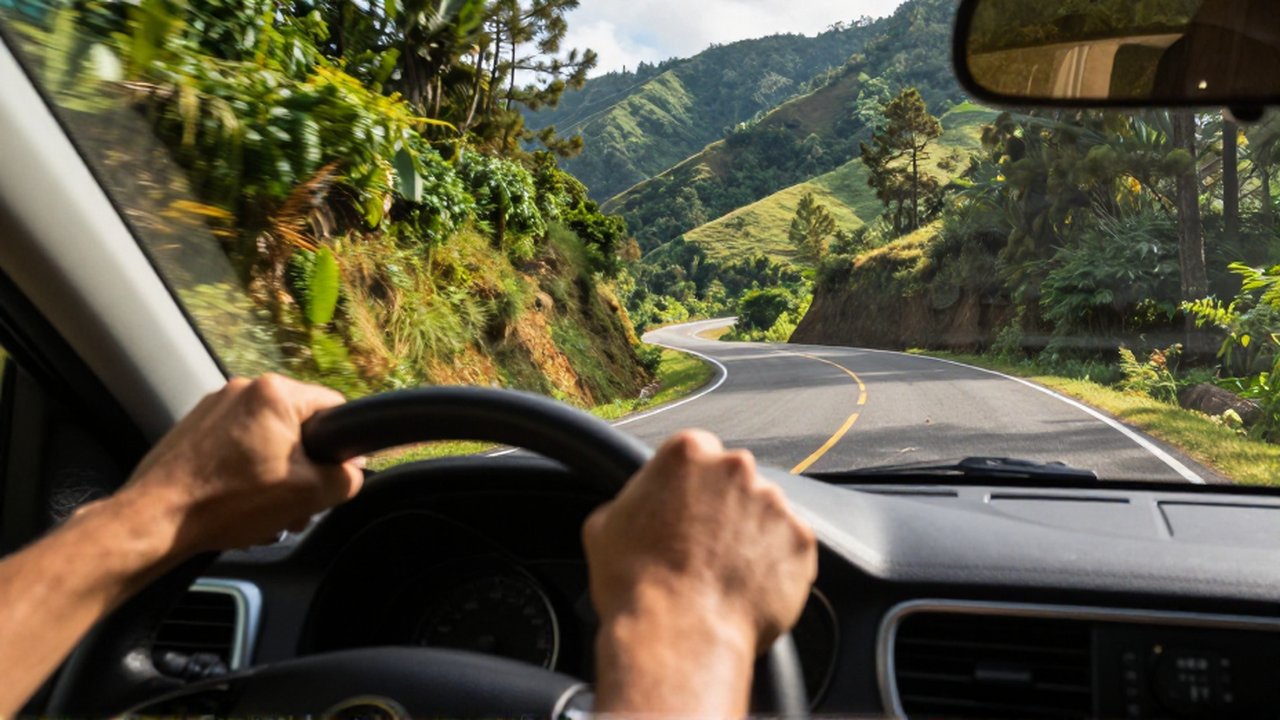 Hands grip a steering wheel while driving an SUV down a winding, sun-drenched mountain road.
